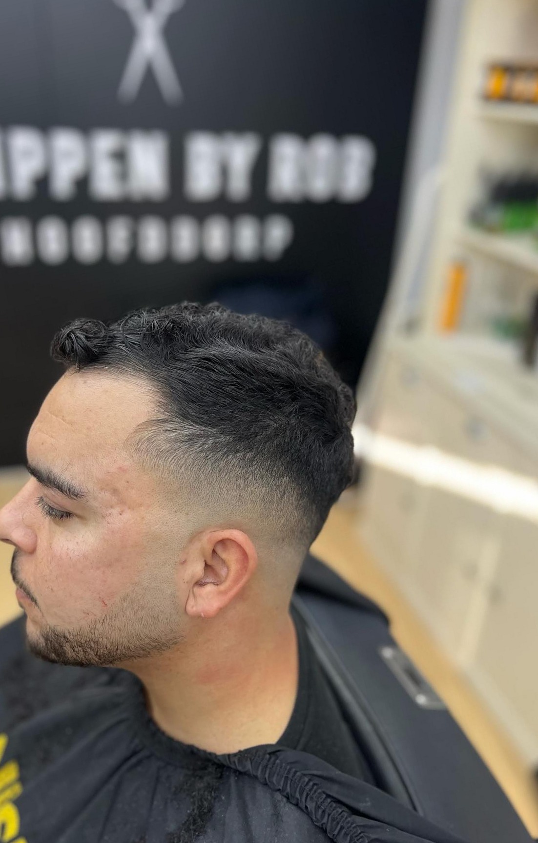 Kort getextureerd kapsel met fade en baard, de muur met de naam van de zaak erachter