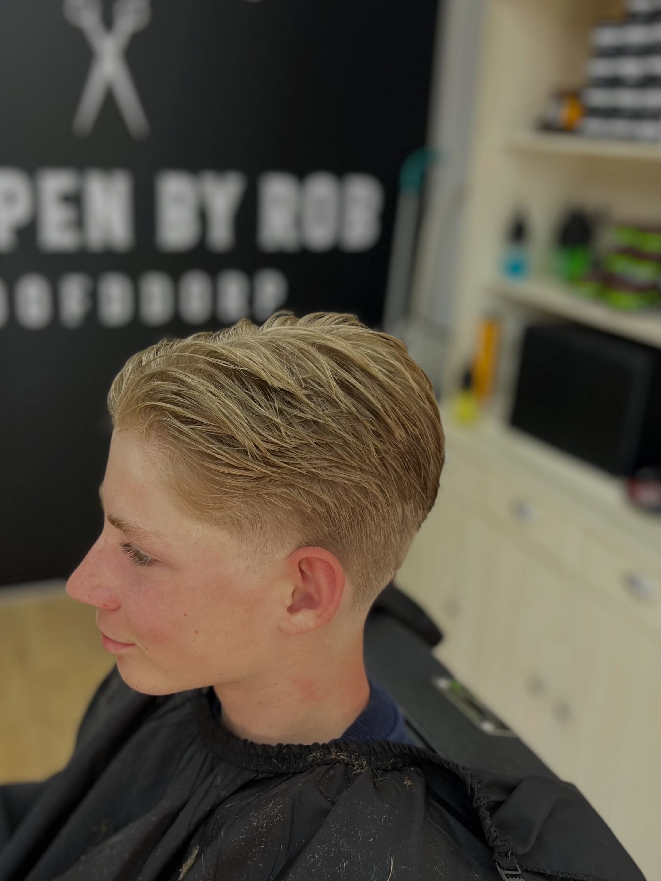Blonde fade van opzij, met de zwarte muur en de letters Knippen by Rob Hoofddorp op de achtergrond